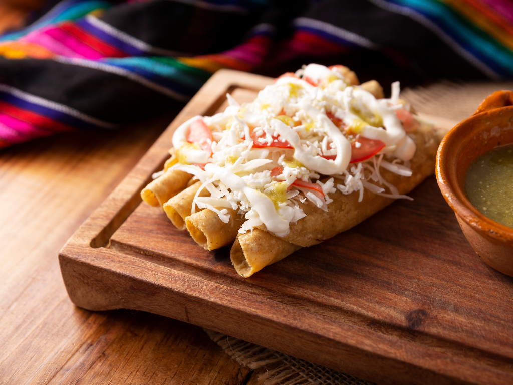 Freshly made authentic Mexican taquitos topped with crema, shredded cheese, lettuce, and tomato. Learn to prepare traditional dishes like this at our cooking classes in Crest Hill, Mokena, and Chicago, Illinois