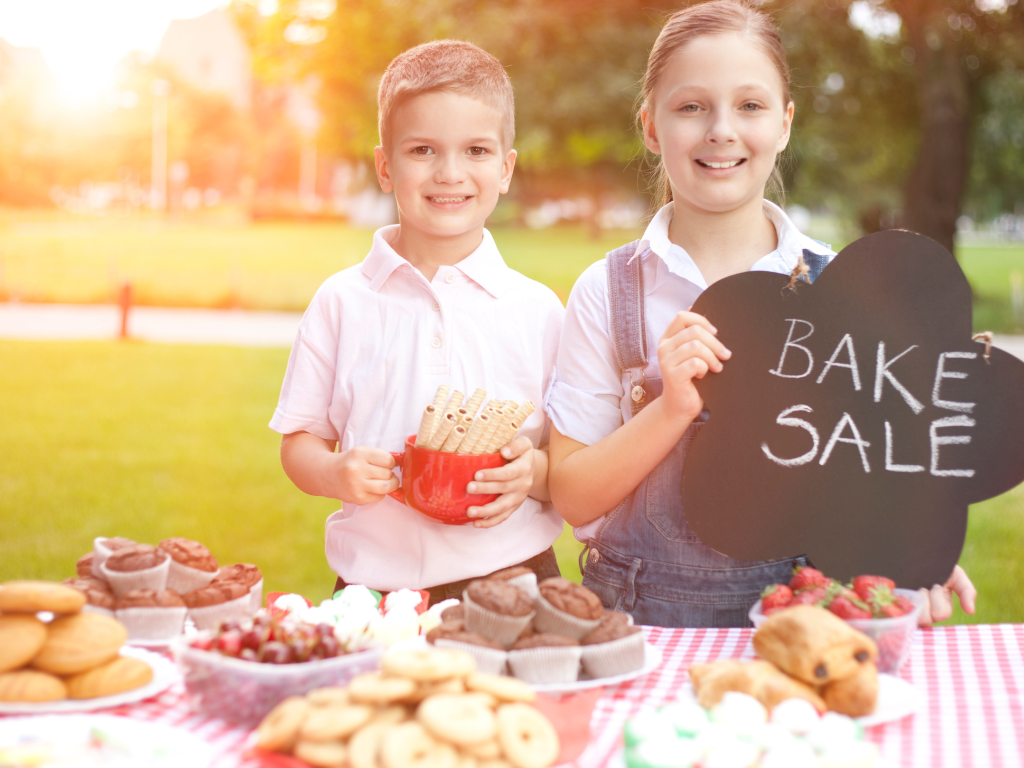 Two children hosting a bake sale table with cookies, muffins, and treats after a kids cooking event at Farmhouse Academy near Plainfield, Shorewood, and Romeoville, Illinois