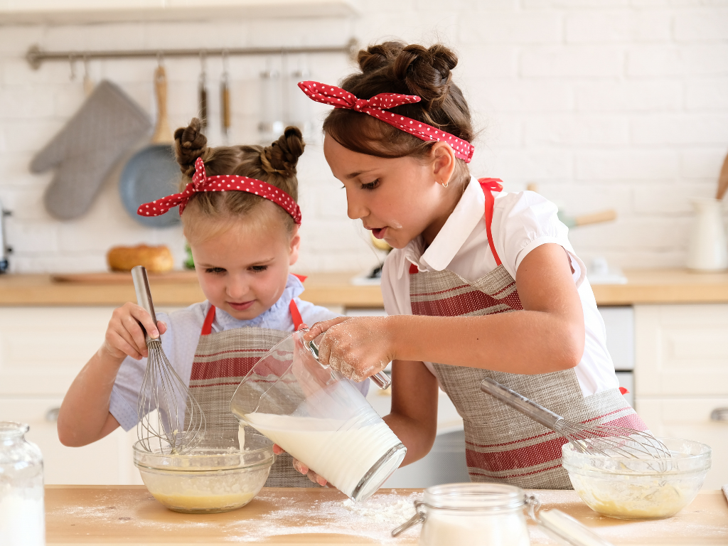 Two young girls in aprons baking together, pouring milk and whisking batter in a kids' baking class in Chicago, Crest Hill, or Mokena.