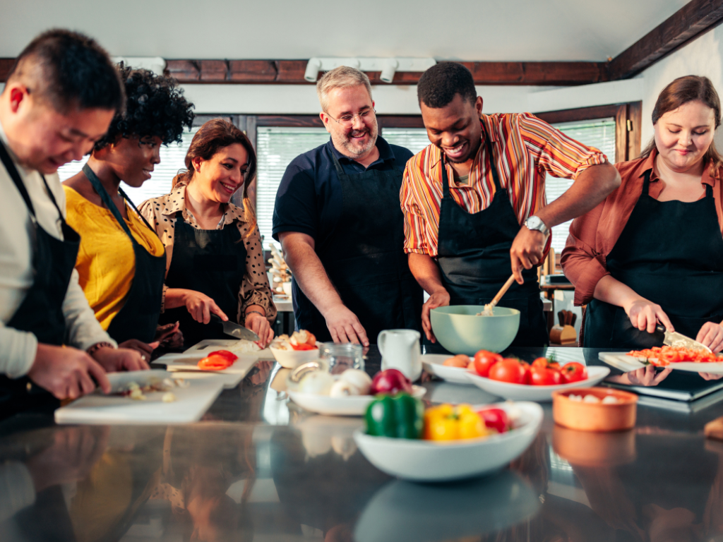 Group of diverse adults smiling and cooking together during a hands-on cooking class with fresh ingredients on the table.