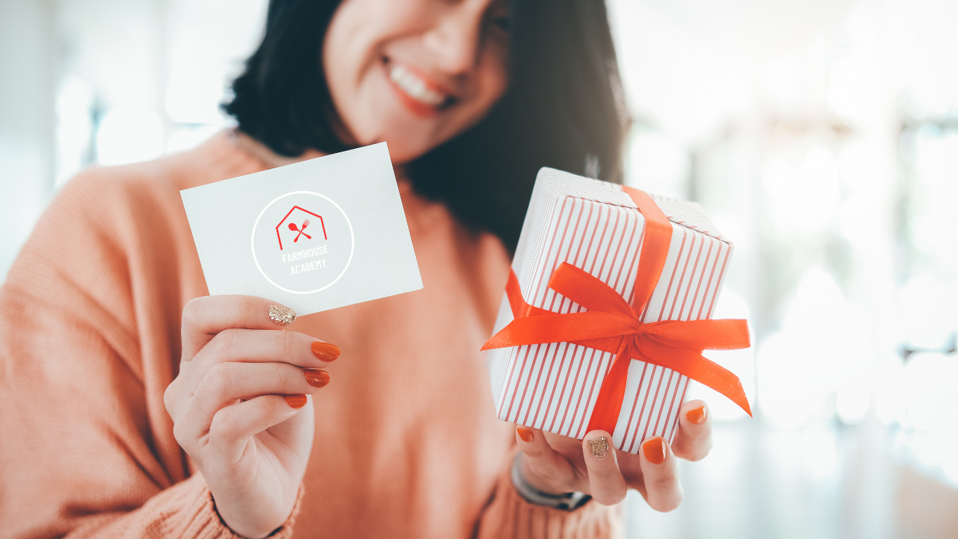 Smiling woman holding a Farmhouse Academy gift card and wrapped present, perfect for gifting cooking classes, baking camps, and culinary experiences in Mokena, Illinois