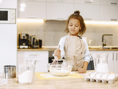A young girl in a beige apron joyfully whisks eggs and flour in a modern kitchen, capturing the warmth and creativity of home baking. Perfect for family cooking blogs or lifestyle features.