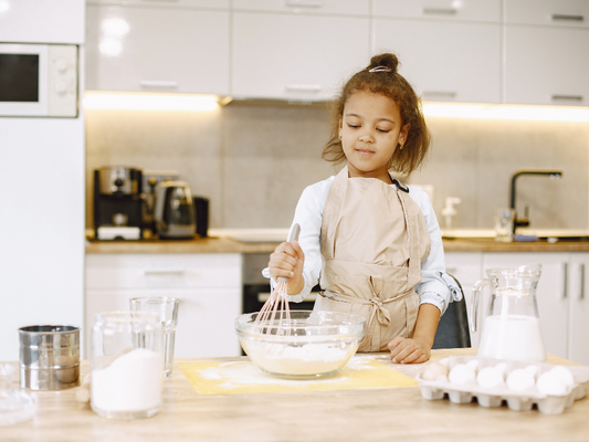 A young girl in a beige apron joyfully whisks eggs and flour in a modern kitchen, capturing the warmth and creativity of home baking. Perfect for family cooking blogs or lifestyle features.