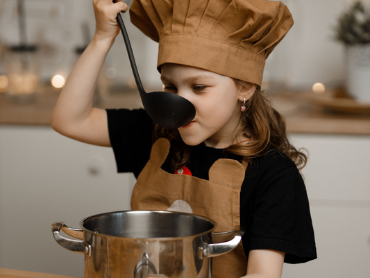 A young girl dressed in a brown chef hat and apron tastes soup from a ladle while cooking in a cozy kitchen, capturing the joy and creativity of childhood culinary adventures.